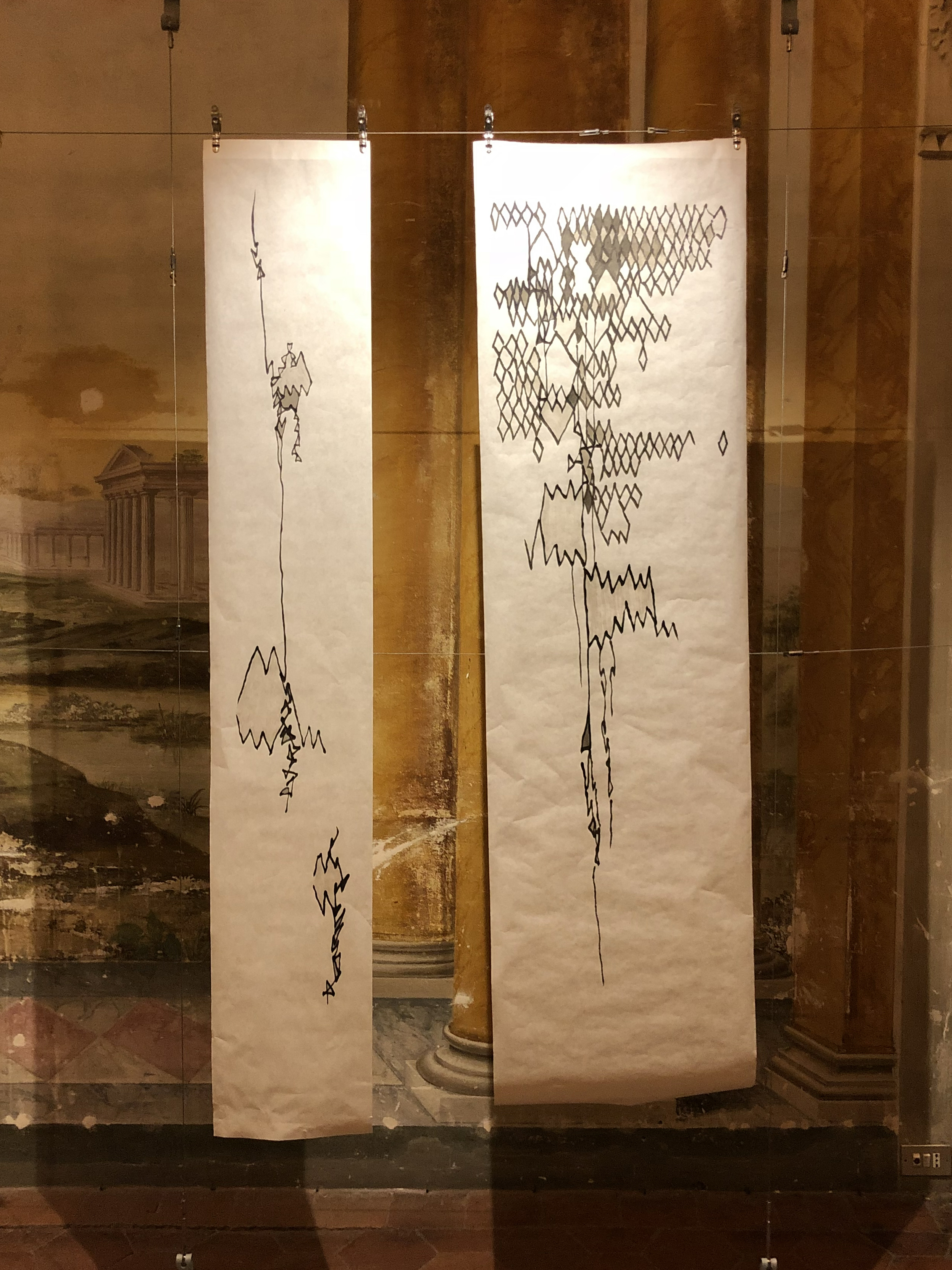 Lines 1 and 2 by Shannon Milar — Life Inflamed series. Ink on paper, 2018. Installation view at Villa Bottini, Lucca. Two tall, narrow vertical sheets hung side by side before the sfumato fresco walls. Left (Lines 1): a sparse, descending vertical mark with small accumulated clusters at intervals — hesitant, fragmented. Right (Lines 2): a dense crosshatched accumulation at the top descends through zigzag and gathered marks toward the base. The architectural painting is visible behind both sheets.