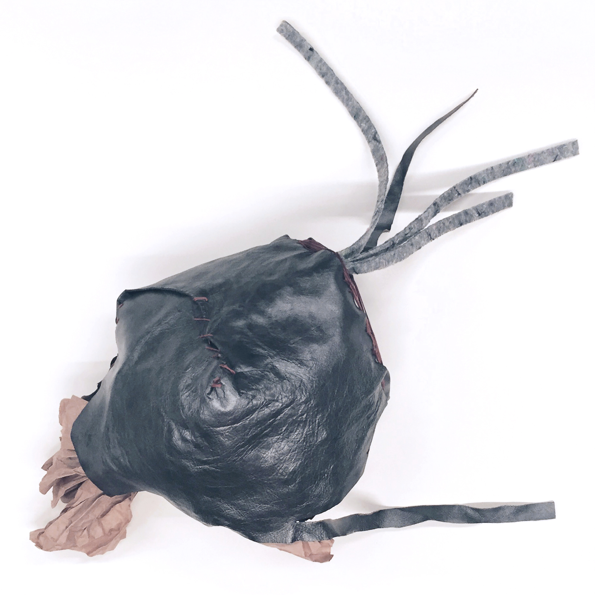 Newcomer by Shannon Milar — Moondream series. A bulbous dark leather form, stitched closed with burgundy embroidery floss in a spiral at its centre, trailing strips of grey weatherproofed material from its crown. Crumpled craft paper at the base.